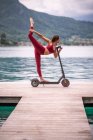Vista lateral da fêmea flexível encantada em balanceamento activewear em Natarajasana em scooter elétrico enquanto pratica ioga no cais de madeira e olhando para longe — Fotografia de Stock