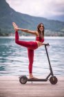 Vista lateral de la mujer flexible encantada en ropa deportiva equilibrio en Natarajasana en scooter eléctrico mientras practica yoga en el muelle de madera y mirando a la cámara - foto de stock