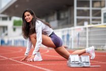 Visão lateral do corpo inteiro de sorrir jovem corredor feminino hispânico em sportswear fazendo exercício lunge enquanto estirando as pernas antes de correr na pista do estádio — Fotografia de Stock