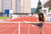 Jovem atleta hispânica do sexo feminino que alonga as pernas perto do corrimão de metal enquanto se aquece antes de correr na pista do estádio — Fotografia de Stock