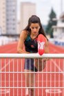 Young Hispanic female athlete stretching legs near metal railing while warming up before running on stadium track — Stock Photo