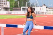 Jovem atleta hispânica positiva em sportswear sentado na barreira e sorrindo enquanto descansa durante o treino na pista de corridas do estádio — Fotografia de Stock