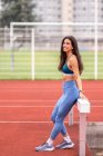 Позитивная молодая латиноамериканка в спортивной одежде сидит на барьере и улыбается во время перерыва на ипподроме стадиона — стоковое фото