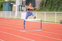 Comprimento total de confiante jovem esportista hispânica pulando sobre a barreira enquanto corre na pista vermelha do estádio de esportes — Fotografia de Stock
