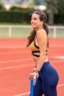 Felice giovane atleta donna ispanica in elegante reggiseno sportivo e leggings guardando altrove mentre in piedi allo stadio — Foto stock