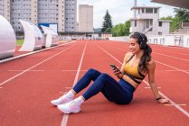 High angle of tired young fit Hispanic sportswoman in activewear and sneakers sitting on red track near smartphone and headphones while resting after workout at stadium — Stock Photo