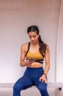 Молодая спортсменка-латиноамериканка в спортивной одежде сидит на скамейке и использует мобильное приложение на смартфоне во время отдыха после тренировки — стоковое фото