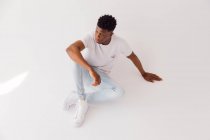 Desde arriba cuerpo completo de joven afroamericano modelo masculino con camiseta blanca con pantalones vaqueros y zapatillas de deporte azul claro sentado en el suelo en el estudio - foto de stock