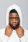 Confident serious young African American male in white hoodie with hood on head looking at camera against gray background — Stock Photo