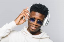 Joven chico hipster afroamericano feliz con capucha blanca y gafas de sol de moda escuchando música a través de auriculares inalámbricos - foto de stock