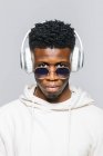 Happy young African American hipster guy in white hoodie and trendy sunglasses listening to music through wireless headphones — Stock Photo