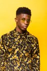 Portrait of unemotional African American male in trendy outfit standing on yellow background in studio looking at camera — Stock Photo