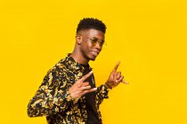 Smiling African American male in trendy outfit showing shaka gesture on yellow background in studio — Stock Photo