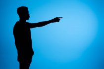 Вид сбоку на силуэт афроамериканского мужчины, стоящего с протянутой рукой и указывающего на синий фон в студии — стоковое фото