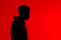 Вид сбоку на силуэт неузнаваемого афроамериканского мужчины в толстовке, стоящего на красном фоне в темной студии — стоковое фото