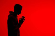 Seitenansicht der Silhouette eines nicht wiederzuerkennenden afroamerikanischen Mannes, der mit gefalteten Händen auf rotem Hintergrund im Studio steht und betet — Stockfoto