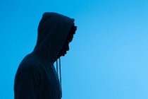 Seitenansicht der Silhouette eines nicht erkennbaren afroamerikanischen Mannes in Kapuzenpulli, der auf blauem Hintergrund im dunklen Studio steht — Stockfoto