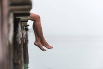 Side view of crop legs of female sitting on wooden quay near sea on misty morning on Playa de Muro — Stock Photo