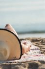 Mujer anónima con sombrero de paja tumbada en la playa de arena por la mañana y tomando el sol durante las vacaciones de verano en Playa de Muro - foto de stock