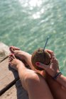 D'en haut de femelle méconnaissable assis avec cocktail de noix de coco avec de la paille près de la mer bleu ondulation et profiter des vacances d'été sur Playa de Muro — Photo de stock