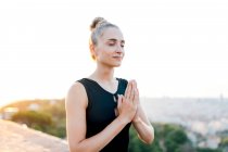 Heiteres Weibchen mit geschlossenen Händen auf der Brust und geschlossenen Augen meditiert auf dem Dach während der Yoga-Praxis am Abend — Stockfoto