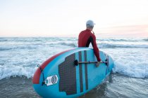 Вид ззаду на невизначений чоловічий серфер у гідрокостюмі та капелюсі, що несе дошку весла та входить у воду для серфінгу на березі моря — стокове фото