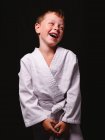 Симпатичний хлопчик у карате кімоно щасливо сміється з відкритим ротом в студії на чорному тлі — стокове фото