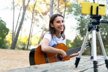 Alegre blogger adolescente tocando la guitarra acústica mientras graba video en el teléfono celular en el trípode en el parque - foto de stock