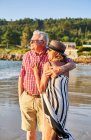Sorrindo descalço casal de idosos em óculos de sol de pé abraçando uns aos outros na praia de areia molhada e desfrutando de dia ensolarado — Fotografia de Stock