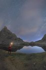 Vista posteriore di un turista maschio irriconoscibile con torcia che ammira monti innevati che si riflettono in acqua sotto il cielo stellato di notte — Foto stock
