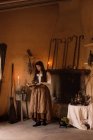 Bruja en vestido largo leyendo el libro mágico de hechizos mientras está de pie en una habitación acogedora con escoba y caldero - foto de stock