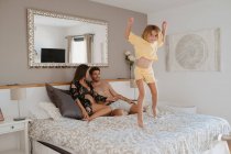 Весела дитина в піжамі розважається на ліжку проти вагітної матері, що взаємодіє з чоловіком в будинку — стокове фото