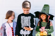 Full body of group of little kids dressed in various Halloween costumes with carved Jack O Lantern browsing mobile phone together while standing near white wall on street — Stock Photo