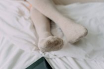 Cortar fêmea irreconhecível em meias de joelho sentado com pernas cruzadas contra celular com tela preta em lençol amassado — Fotografia de Stock