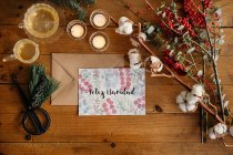 Top view of Christmas composition with colorful postcard with inscription Feliz Navidad placed near burning candles and cups of tea on wooden table decorated with colorful branches of plants — Stock Photo