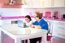 Smiling granddaughter and grandmother sitting at table and using laptop in light room in apartment — Stock Photo