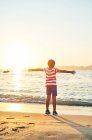Вид ззаду на повну довжину невпізнаваного хлопчика, що стоїть з розтягнутими руками на мокрій піщаному березі, омитому хвилями блакитного моря на заході сонця — стокове фото
