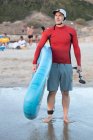 Surfista maschio in muta e cappello in piedi guardando lontano con SUP bordo mentre si prepara a navigare in riva al mare — Foto stock