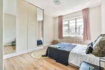 Zeitgenössisches helles Schlafzimmer mit bequemem Bett mit Kissen und Kleiderschrank mit verspiegelter Oberfläche in Fensternähe — Stockfoto