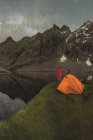 Вид ззаду на невідомий турист, який захоплюється засніженою горою та озером на намет під зоряним небом у сутінках — стокове фото