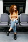 Mujer joven contenta en jeans rasgados con el pelo rojo rizado mirando hacia otro lado en el asiento mientras viaja en tren - foto de stock