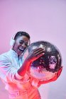 Cool ethnic cheerful woman with short hair in wireless headset and trendy clothes dancing hip hop with open mouth and eyes closed holding glitter ball on violet background — Stock Photo
