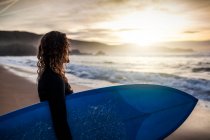 Вид сбоку на неузнаваемую молодую женщину, стоящую на берегу с доской для серфинга, прежде чем попасть в море во время заката на пляже в Астурии, Испания — стоковое фото