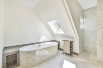 Ванна з мозаїчною плиткою проти стійки з рушниками та вікнами у сучасній ванній в сонячний день — стокове фото