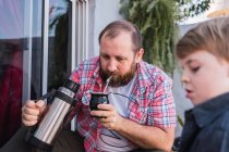 Hipster-Papa gießt Kräutertee aus Thermoskanne und trinkt in Kalebassen-Kürbis gegen Jungen mit Bauholz auf Promenade — Stockfoto