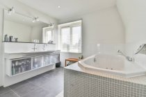 Interior do banheiro contemporâneo com banheira contra cabine de chuveiro e produtos de beleza na prateleira em casa de luz — Fotografia de Stock