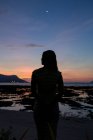 Вид сзади силуэт неузнаваемой женщины-путешественницы, стоящей на песчаном побережье, покрытом водорослями и любующейся закатом в Малайзии — стоковое фото