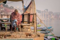 INDIA, VARANASI - 27 DE NOVIEMBRE DE 2015: Varón étnico sentado en un banco de madera cerca de un bote en el dique de un río en la India - foto de stock