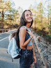 Vista laterale di felice viaggiatore femminile con zaino a guardare la fotocamera mentre si cammina su strada asfaltata a Tenerife Isole Canarie Spagna — Foto stock