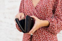 Crop unrecognizable woman in red dress standing and opening black leather purse against light wall in daytime — Stock Photo